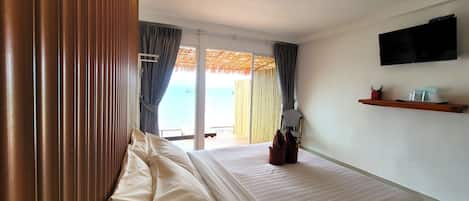 A hotel room with a bed, a TV mounted on the wall, a small shelf with items, and a view of the sea through the window.