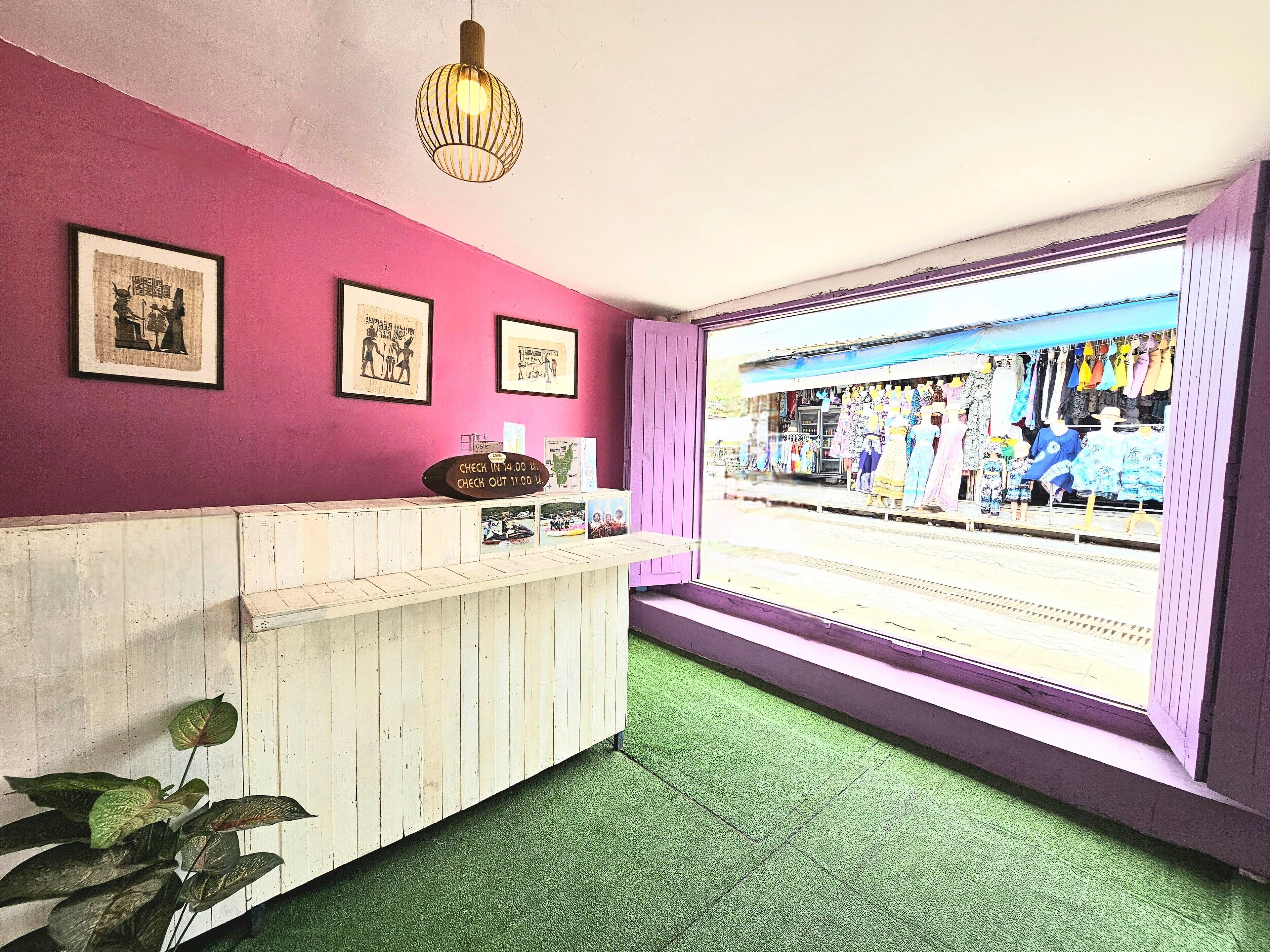 A reception area with a pink wall, framed pictures, a wooden counter, and a view of a clothing store through a window.