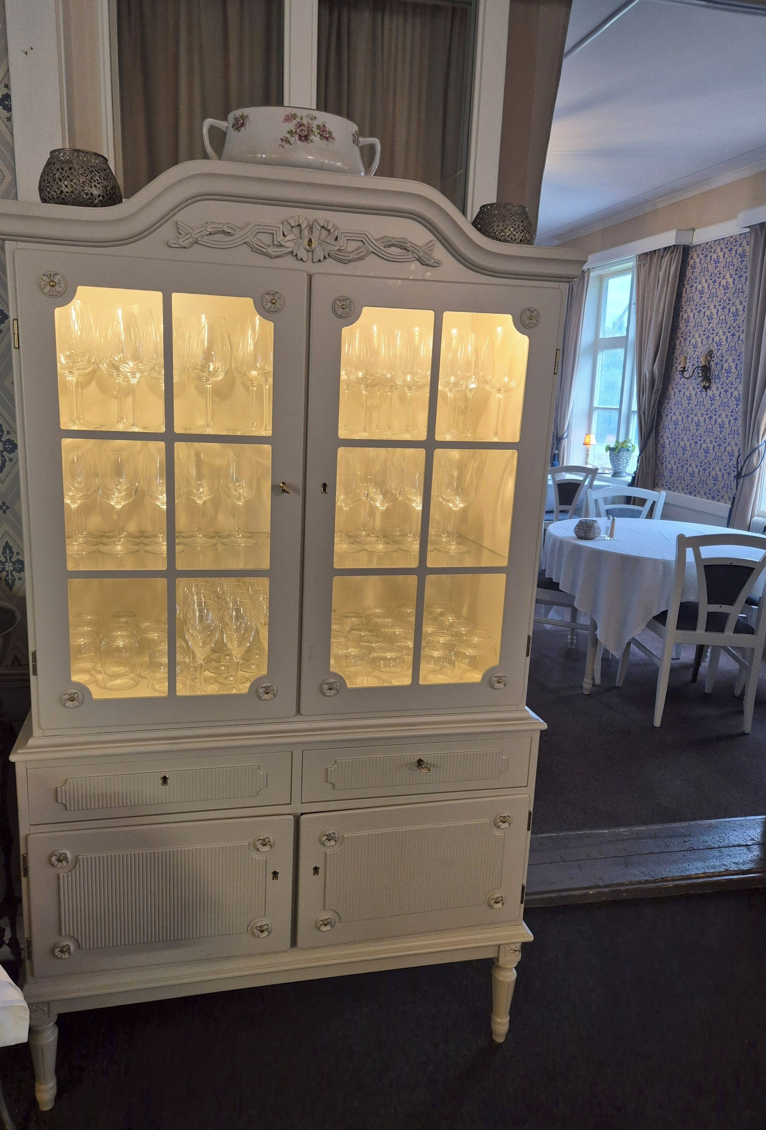 Un mueble blanco y ornamentado con puertas de cristal y cajones, ubicado en una habitación con comedor y papel tapiz estampado.