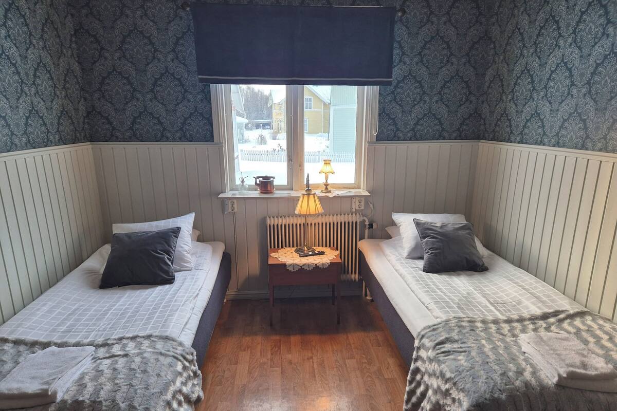Una habitación pequeña y acogedora con dos camas individuales, una mesita con una lámpara y una ventana que da a vistas de casas.