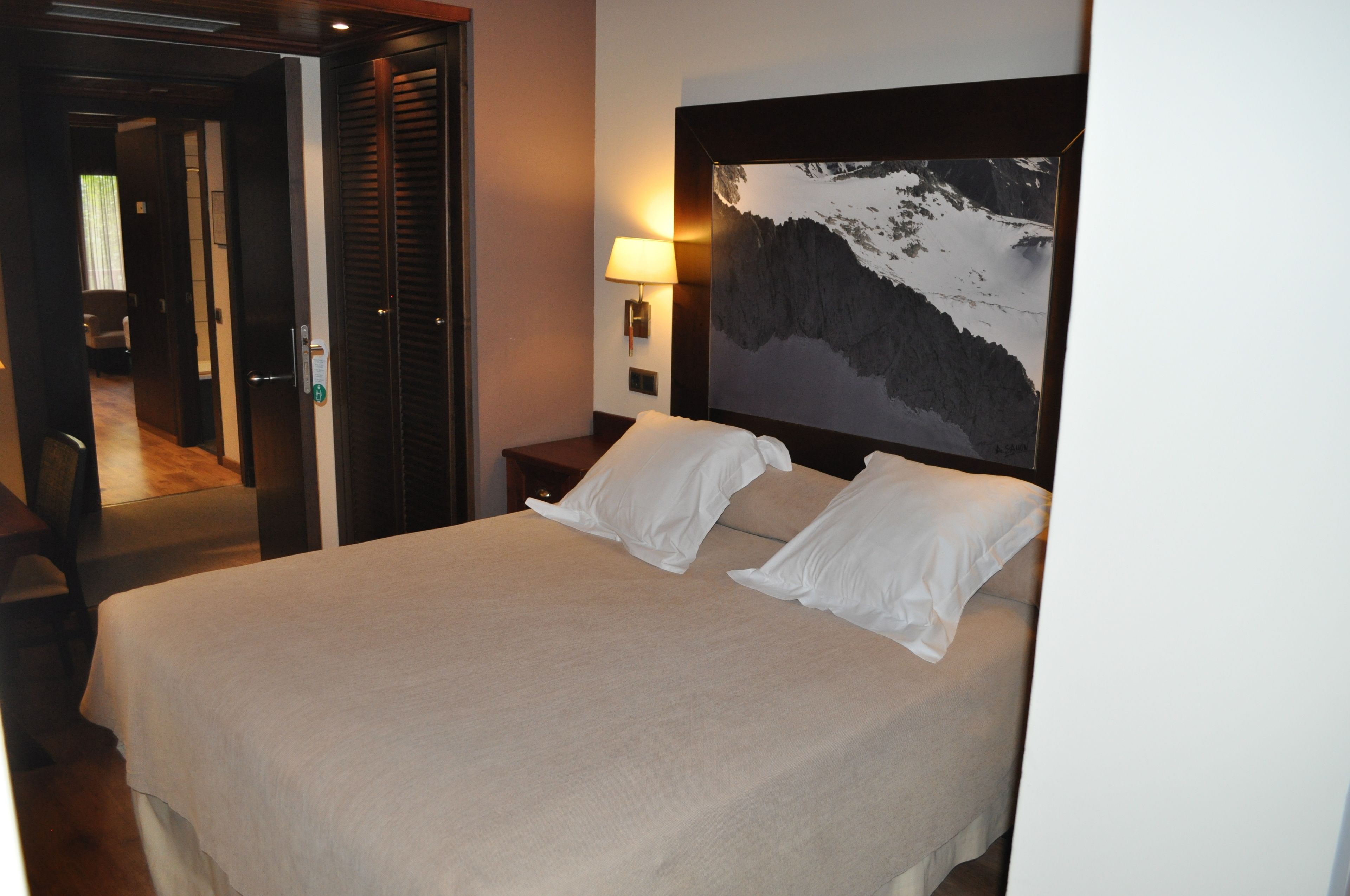 Una habitación de hotel con una cama, una mesita de noche con lámpara y un cuadro grande de un paisaje montañoso.