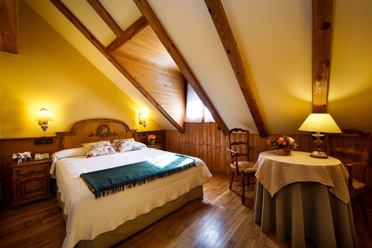 A cozy bedroom with a wooden ceiling, a large bed with a quilt, bedside lamps, a chair, and a table with a lamp.