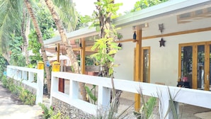 A building with a white exterior, large windows, and a wooden deck. There are plants and a sign indicating a room type.