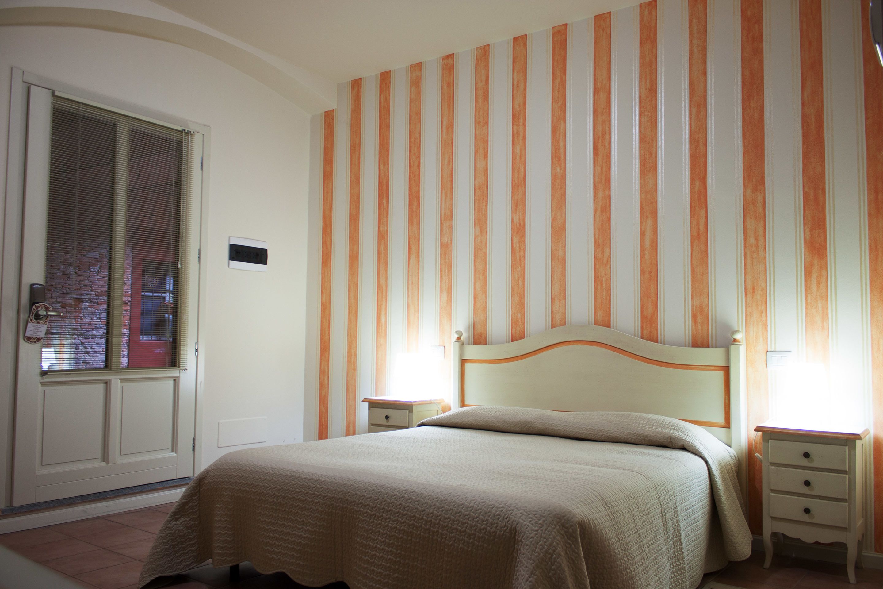 Un dormitorio con una cama, dos mesitas de noche, una puerta con ventana y papel pintado a rayas.