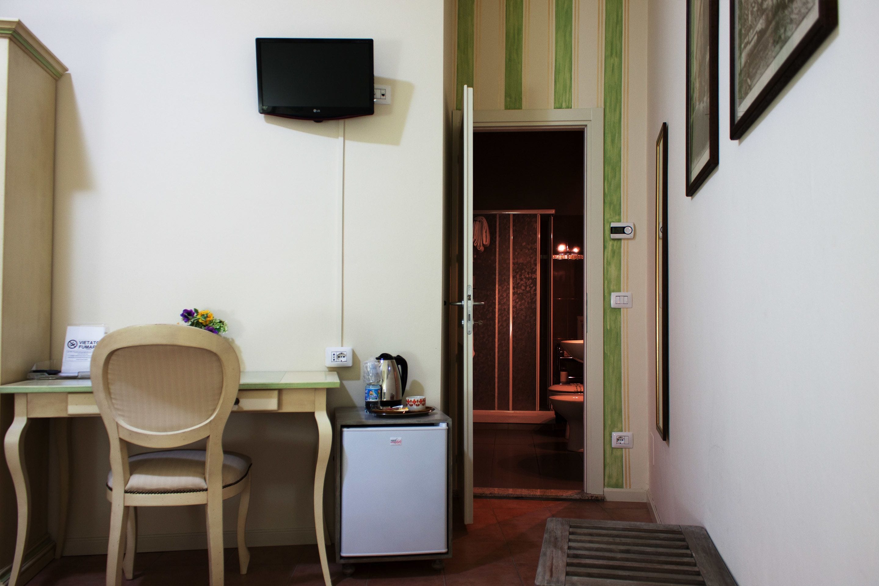 A compact room with a desk, chair, a small refrigerator, a television mounted on the wall, and a door leading to another room.