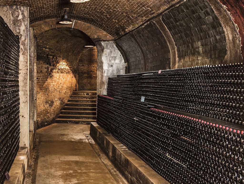 A wine cellar with rows of wine bottles, a brick wall, and a staircase leading to a higher level.