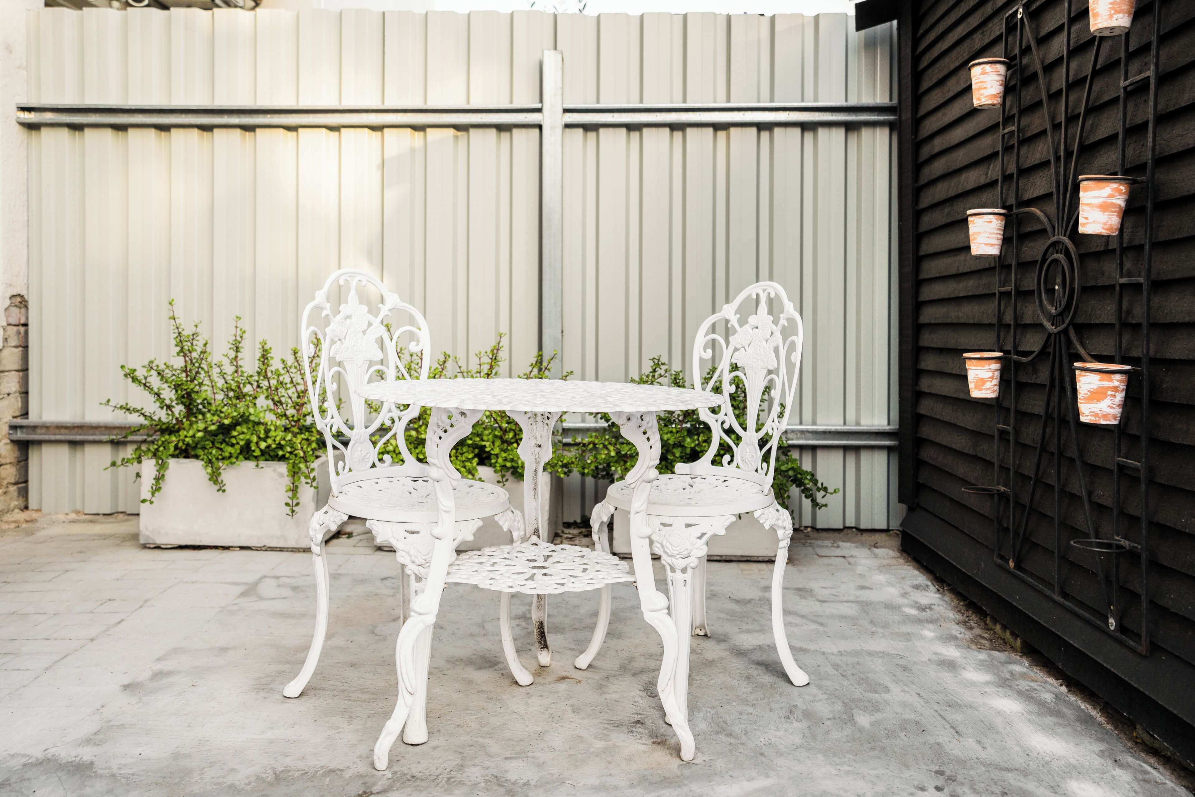 Une terrasse extérieure blanche, avec une table et des chaises, un jardinier contenant des plantes vertes et un mur noir orné de pots suspendus.