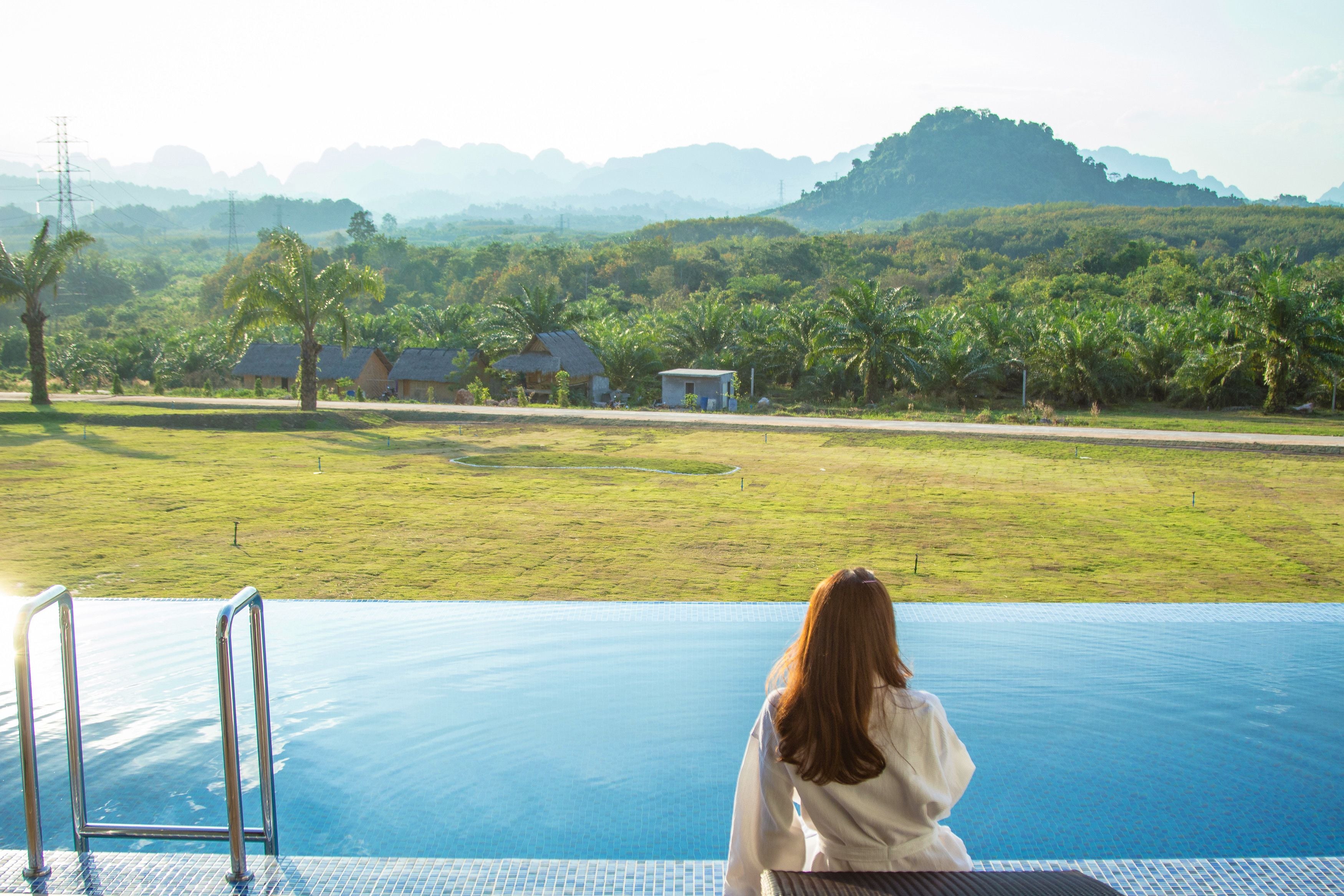 A woman sitting by an infinity pool with a view of a lush landscape and distant mountains.