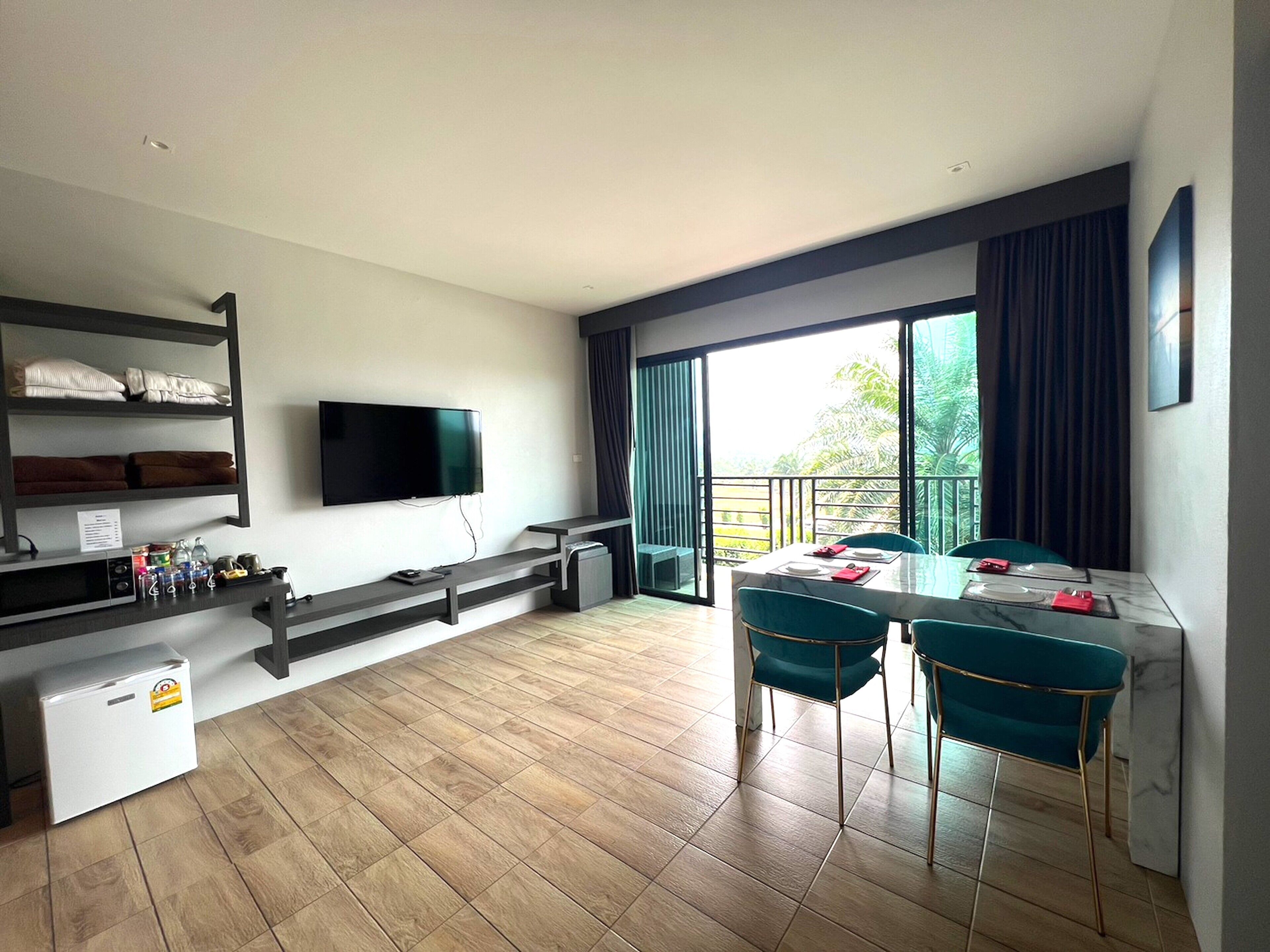 A modern room with a flat-screen TV, a dining table with chairs, a small refrigerator, and a shelf with folded towels.