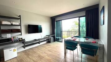 A modern room with a flat-screen TV, a dining table with chairs, a small refrigerator, and a shelf with folded towels.