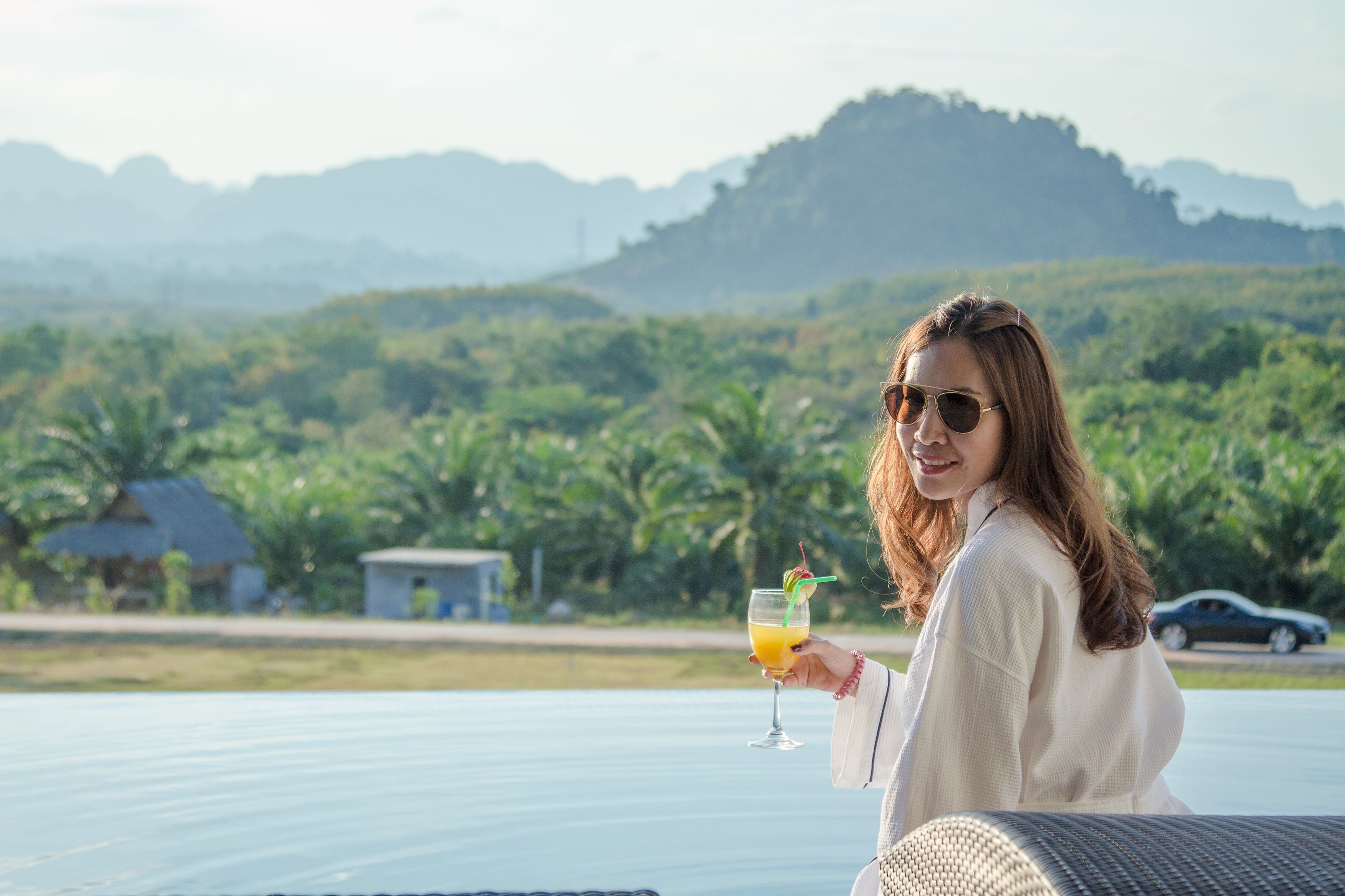 A person holding a drink on a balcony with a view of a lush landscape and mountains in the background.