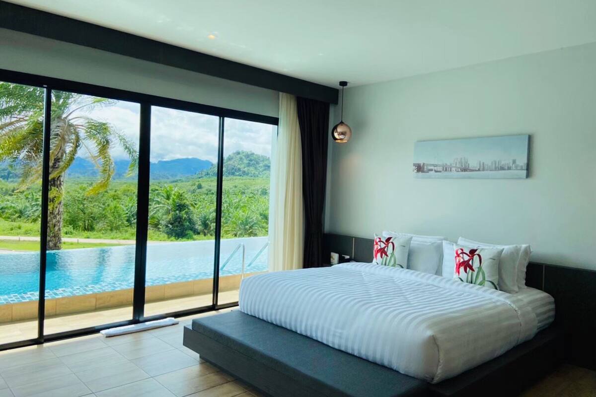 A modern bedroom with a large bed, a cityscape painting, and a view of a lush landscape through floor-to-ceiling windows.