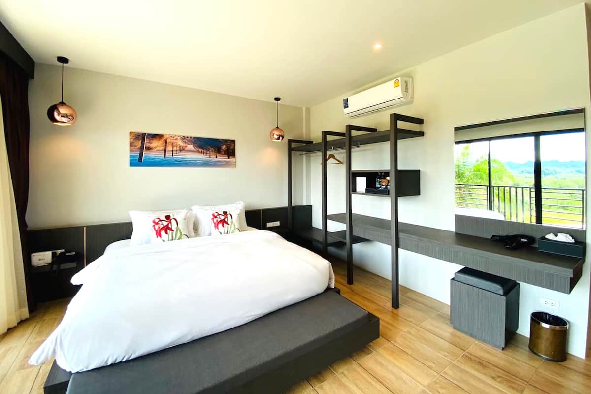 A modern bedroom with a large bed, a desk with a TV, a balcony with a view, and an air conditioning unit.