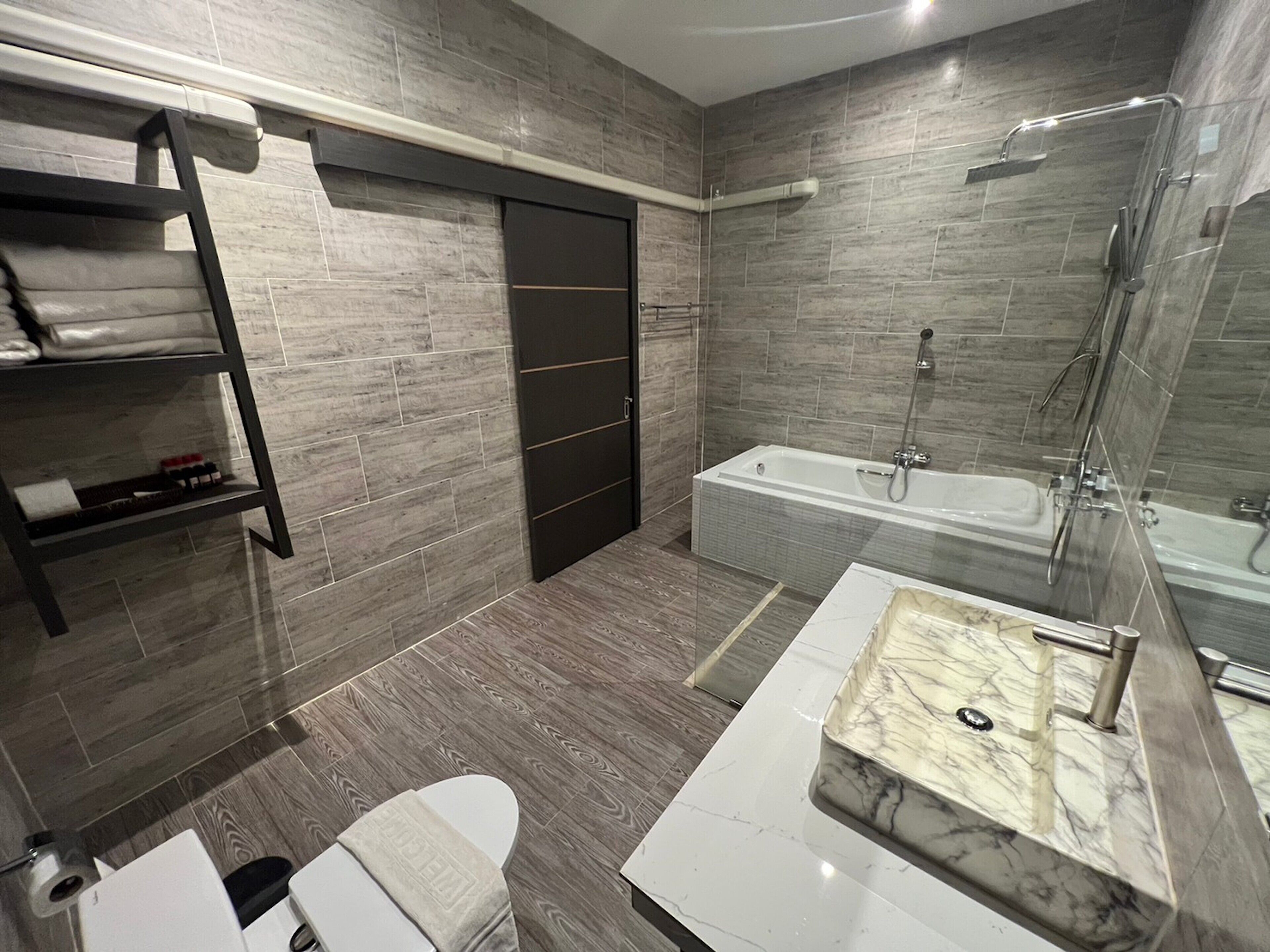 Une salle de bain moderne dotée d’une douche à l’italienne, d’une baignoire, d’un meuble-lavabo avec un plan de travail en marbre et d’une étagère pour les articles de toilette.