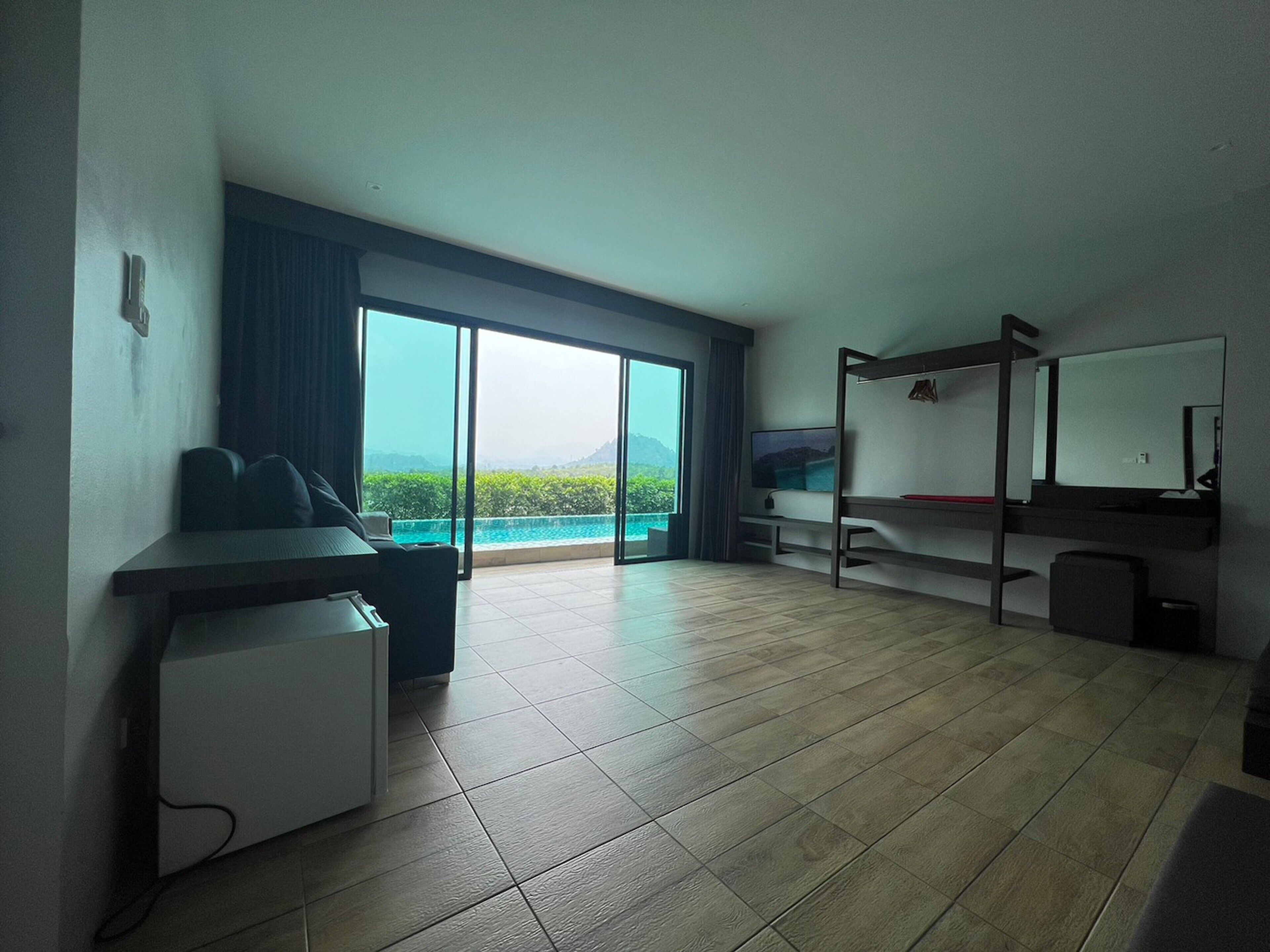 A modern hotel room with a large sliding glass door leading to a balcony with a view of a body of water and mountains. There is a bed, a desk, and a small refrigerator.