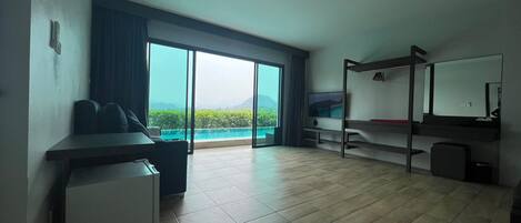 A modern hotel room with a large sliding glass door leading to a balcony with a view of a body of water and mountains. There is a bed, a desk, and a small refrigerator.