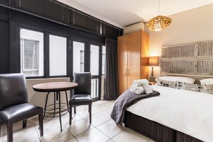 Quarto de hotel com cama, mesa redonda, duas cadeiras, guarda-roupa e uma janela grande.