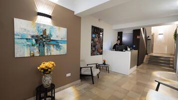 A hotel lobby with a reception desk, a large abstract painting, a vase with flowers, and a staircase leading to another floor.