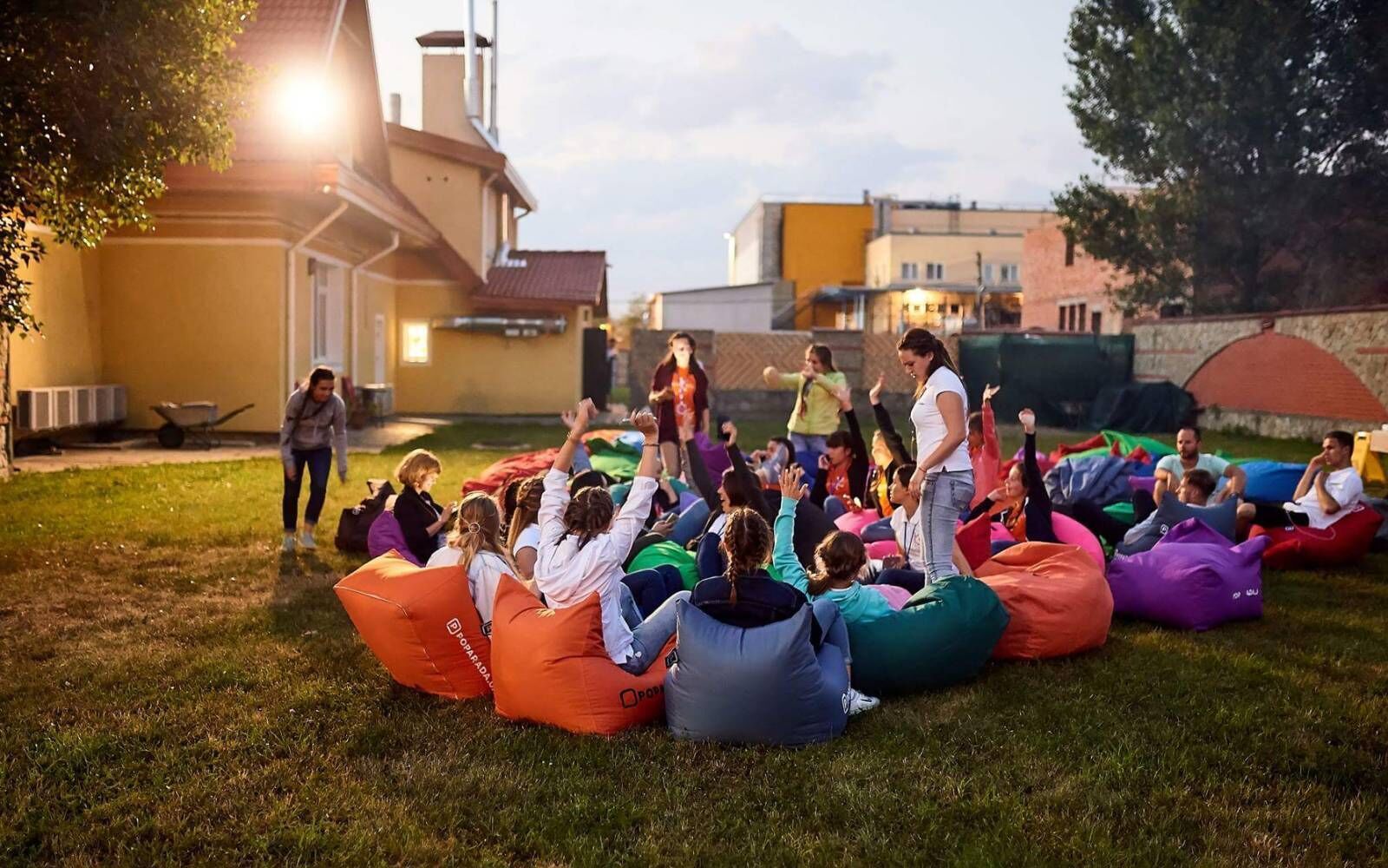 Un groupe de personnes assises sur des poufs colorés, à l’extérieur, au crépuscule.