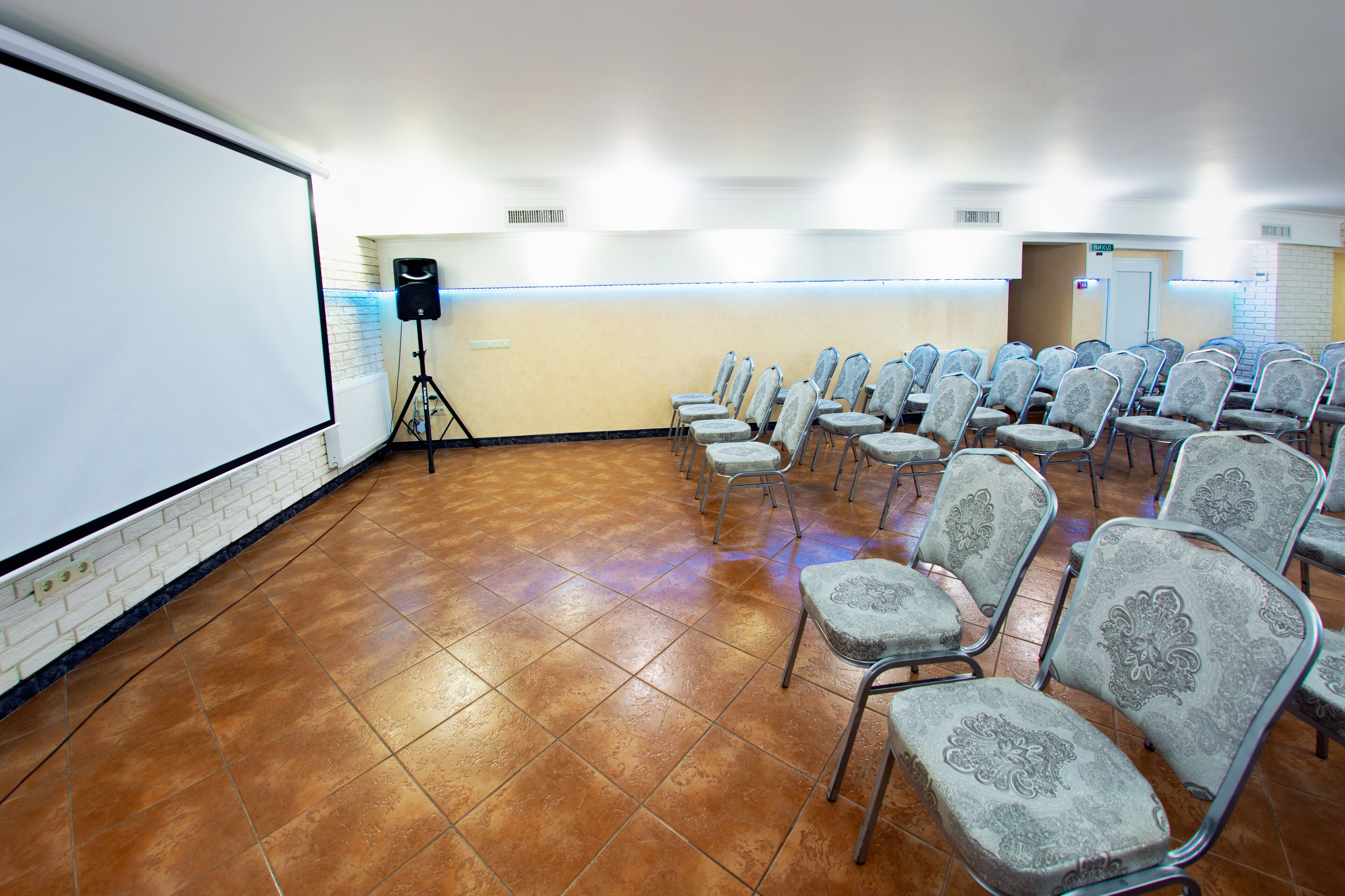 A conference room with a white projection screen, rows of patterned chairs, and a speaker on a stand.