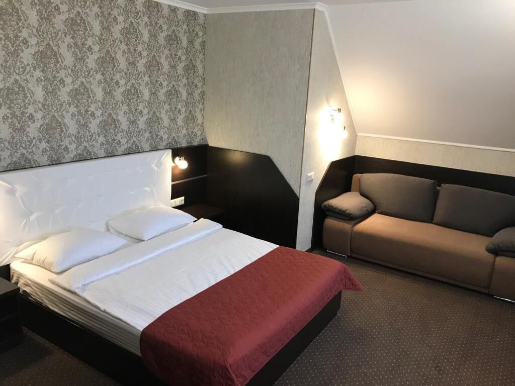 A hotel room with a bed, a red blanket, a brown sofa, and a patterned wallpaper.