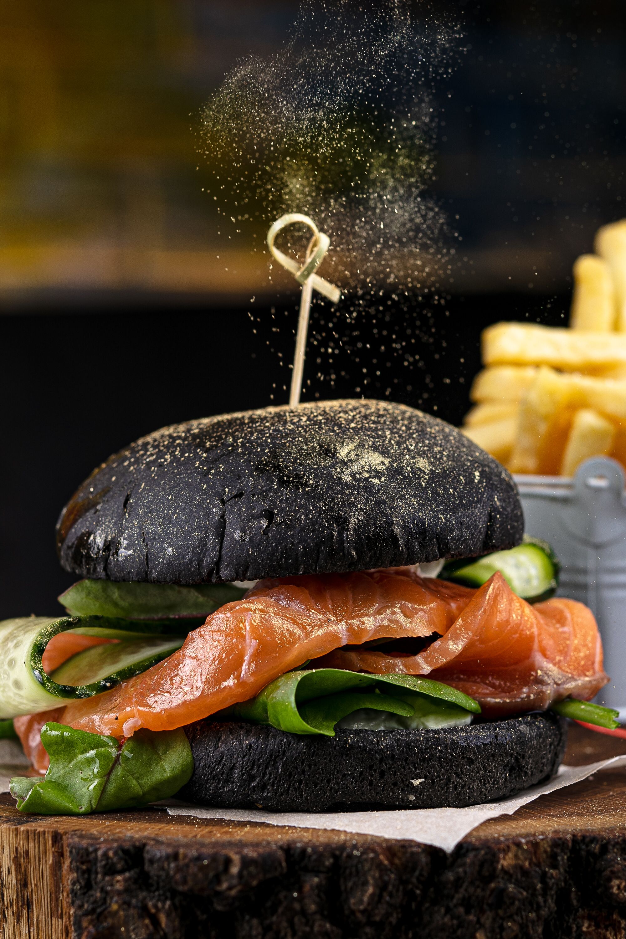 A sandwich with a black bun, salmon, cucumber, and greens, with a side of fries.