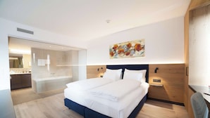 Habitación de hotel moderna con una cama grande, zona de ducha con paredes de vidrio y cabecero de madera.
