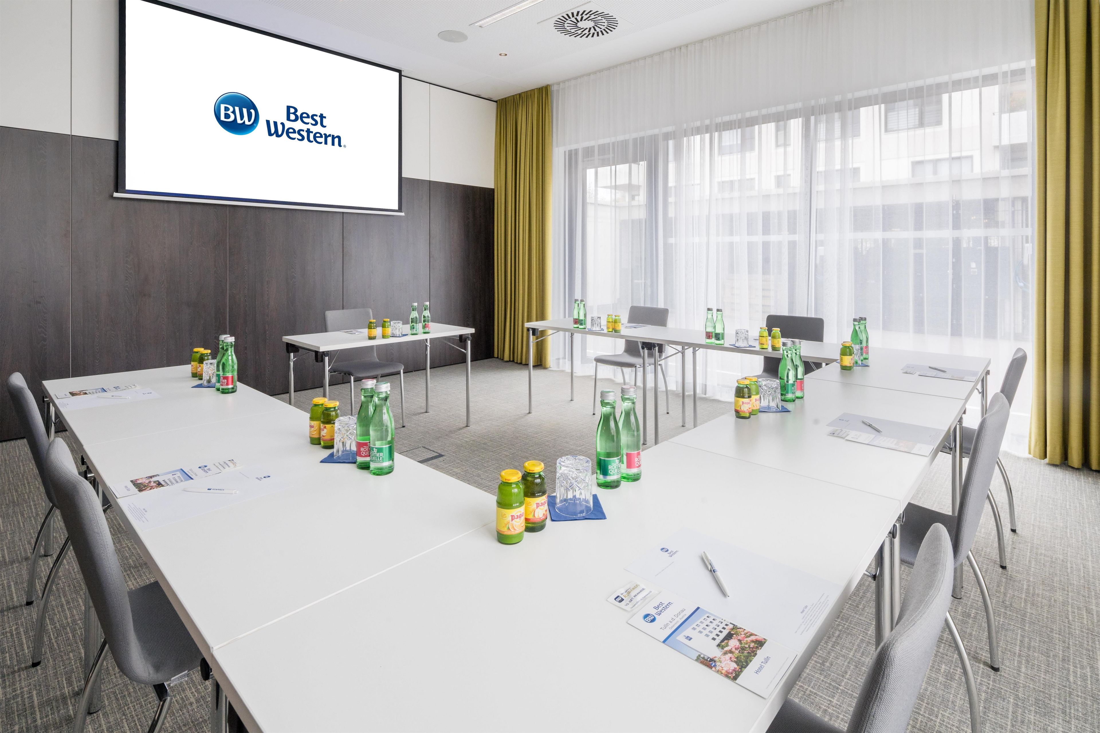 A conference room with white tables, chairs, and a projector screen displaying 'BW Best Western.'