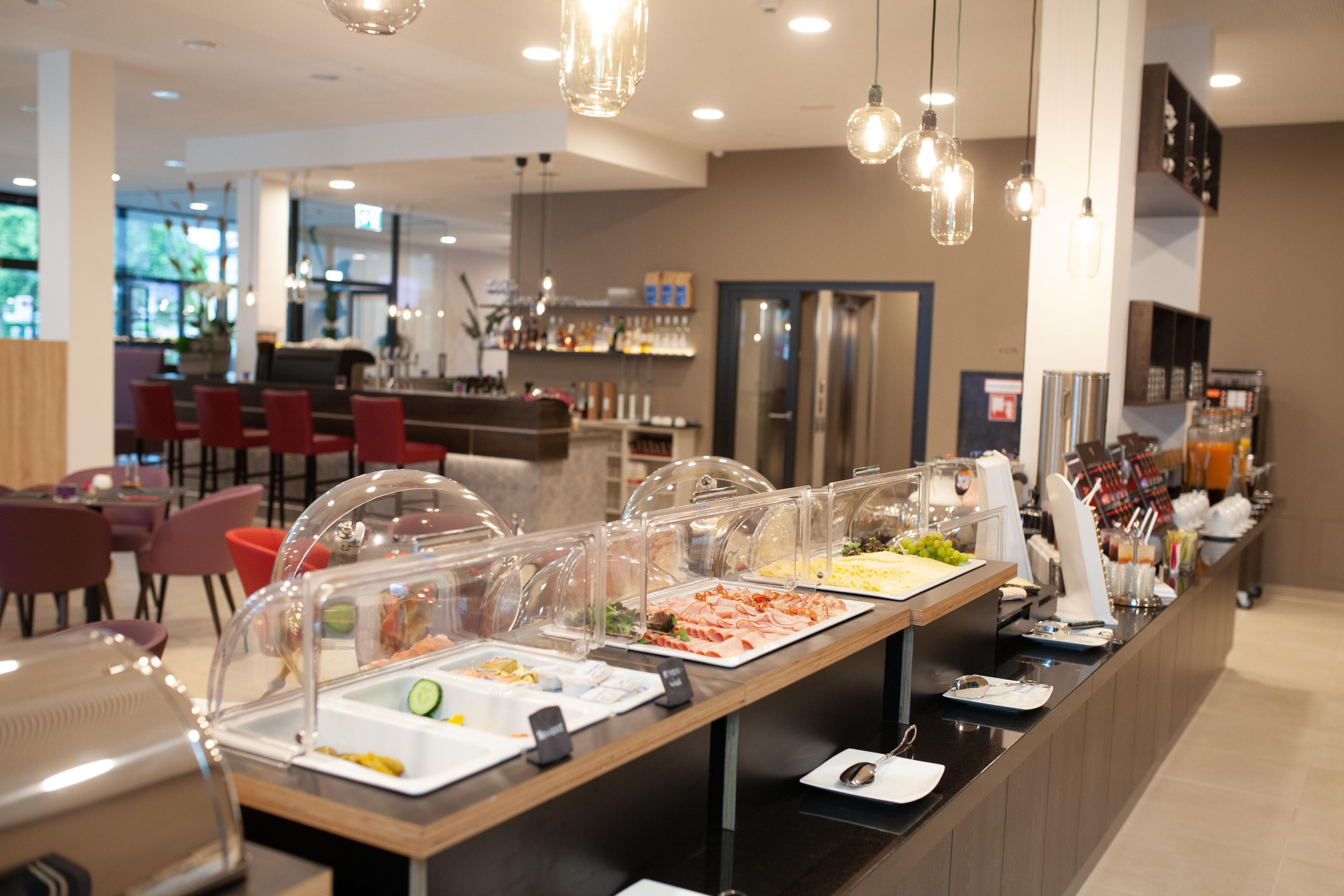 Een buffetgedeelte met diverse gerechten, een zithoek met rode stoelen, en een bar op de achtergrond.