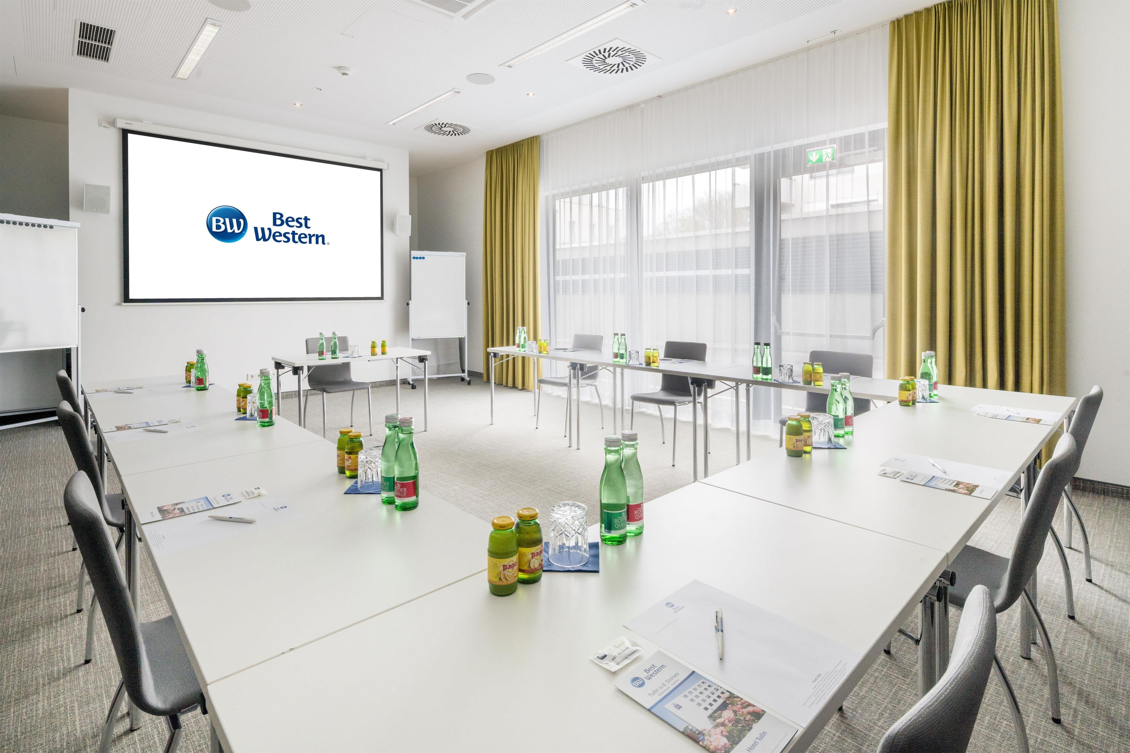 A conference room with a large screen displaying 'Best Western' logo, multiple tables with chairs, and various beverage bottles.