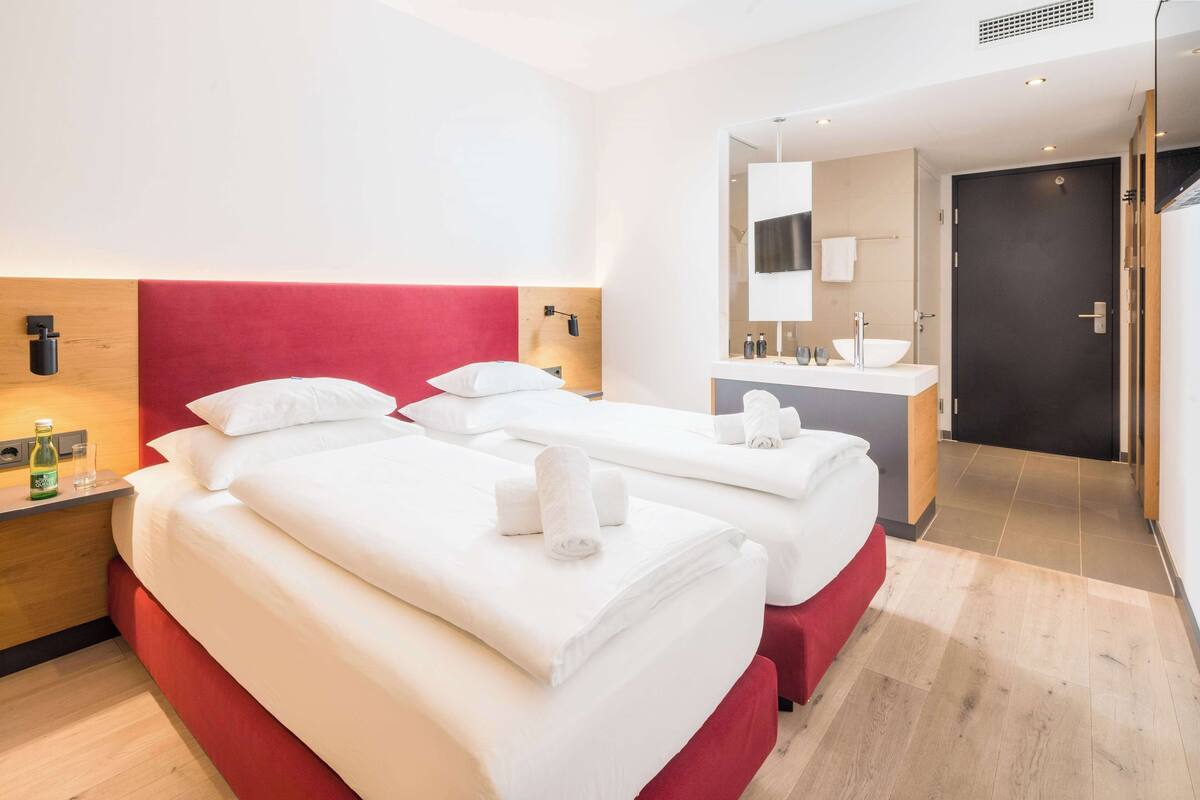 Habitación de hotel con dos camas, una mesita de noche de madera, un espejo, un lavamanos y una puerta.