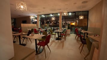 Un intérieur de restaurant moderne, avec de nombreuses tables et chaises, de grandes fenêtres et des luminaires au plafond aux formes géométriques.