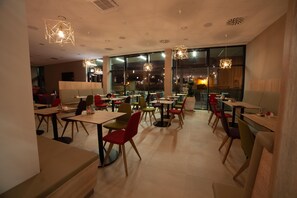 A modern restaurant interior with multiple dining tables and chairs, large windows, and geometric ceiling lights.
