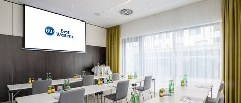 Una sala de conferencias con una pantalla grande mostrando el logotipo de Best Western, varias mesas con sillas y diversas bebidas sobre las mesas.