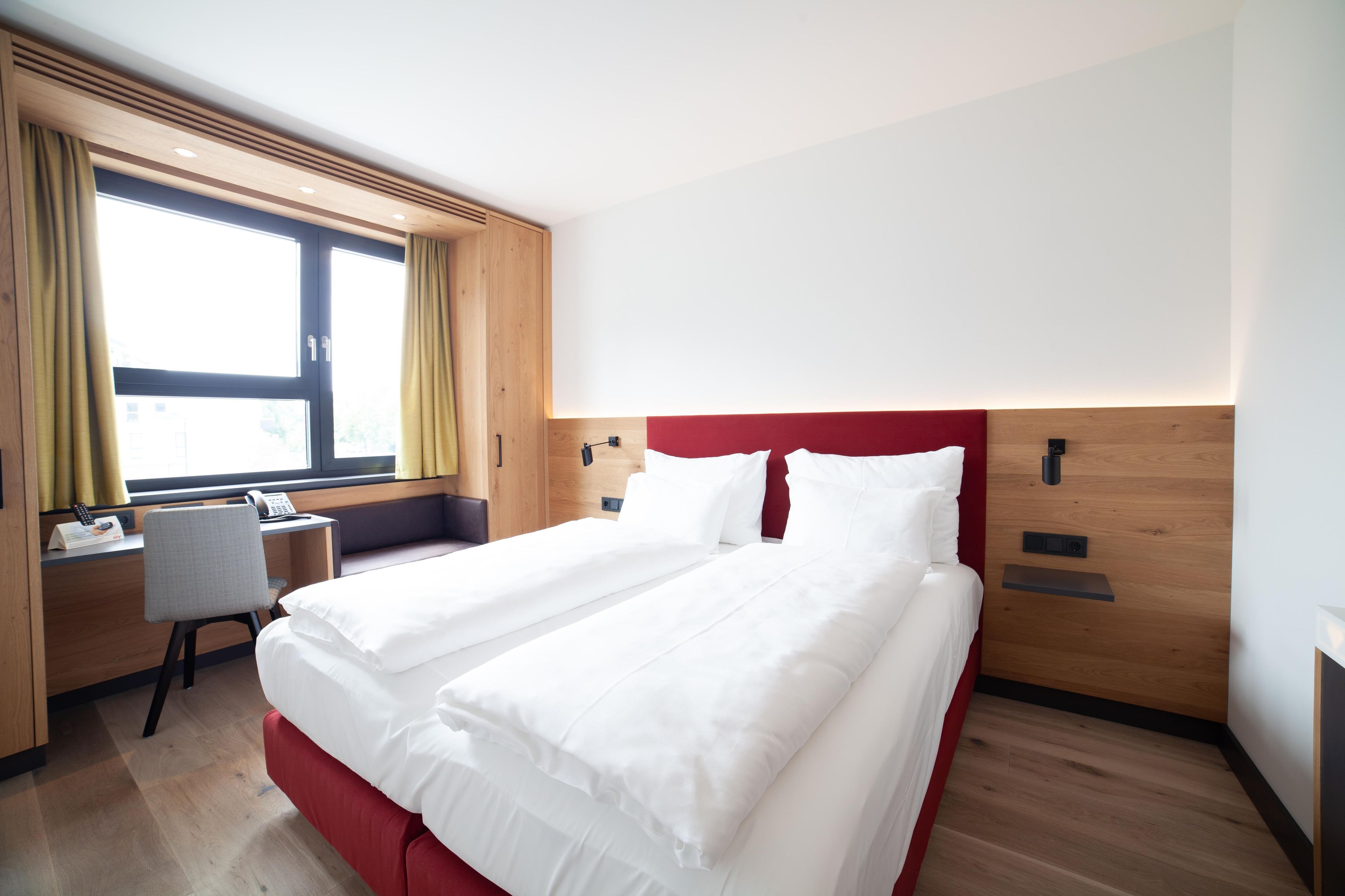 A hotel room with a double bed, a desk with a chair, a window with curtains, and a wooden headboard.