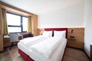 Een hotelkamer met een tweepersoonsbed, een bureau met stoel, een raam met gordijnen en een houten hoofdbord.
