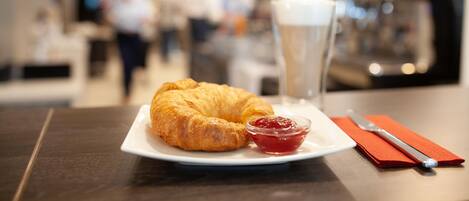 A croissant on a plate with a small bowl of jam and a glass of milk on a table in a cafe setting.
