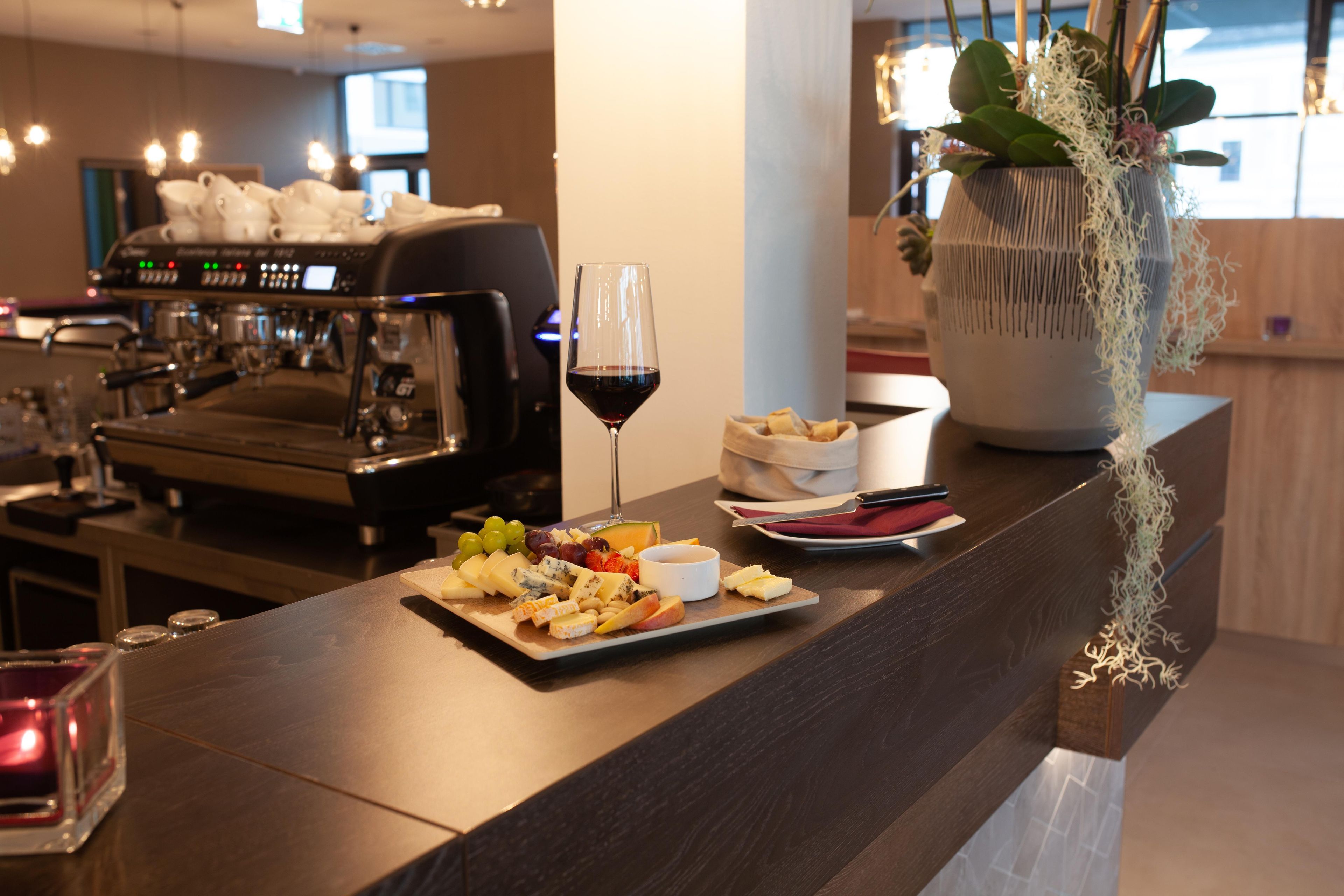 A bar area with a coffee machine, a glass of red wine, and a cheese board with various cheeses and fruits.