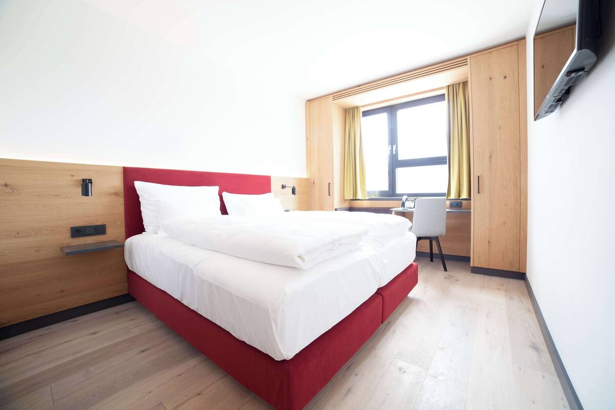 Una habitación de hotel moderna con una cama roja, cabecera de madera y un escritorio con silla.
