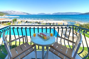 A balcony with a pool, a table with a fruit platter and drinks, and a view of the sea and mountains. - Sami Beach Hotel (Kefalonia)