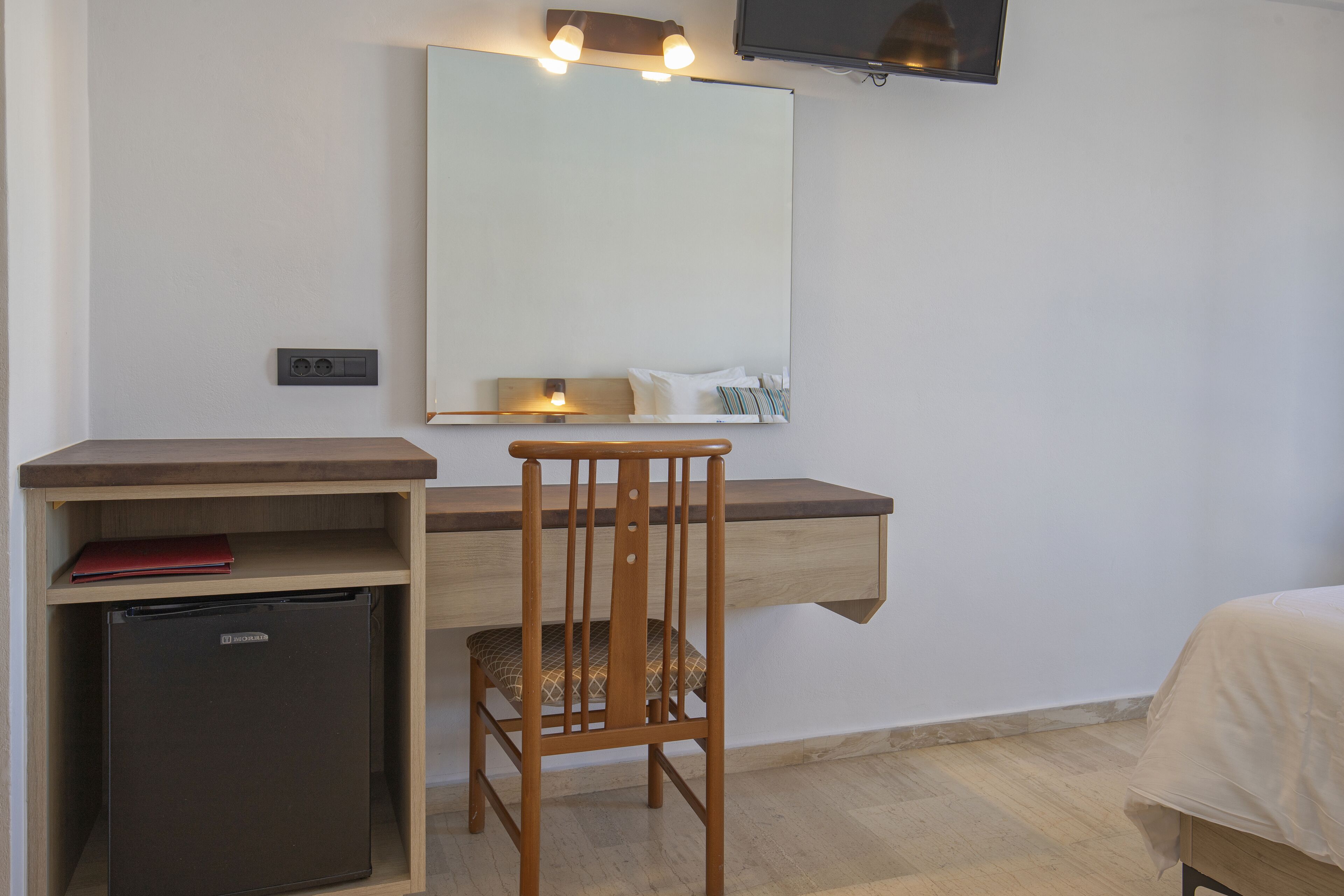 Une chambre d’hôtel compacte comprenant un bureau en bois, un petit réfrigérateur, une chaise, un lit, une table de chevet, un miroir et un téléviseur fixé au mur.