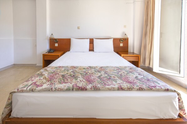 A hotel room with a wooden bed, white bedding, a floral bedspread, two bedside tables with lamps, and a window with curtains. - Sami Beach Hotel (Kefalonia)