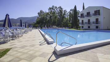 Una piscina con tumbonas y sombrillas, rodeada de un edificio con balcones y vistas a árboles y montañas.