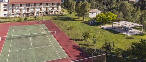 Un court de tennis dont la surface est rouge et verte, entouré d’une clôture. On aperçoit un bâtiment avec de nombreux balcons et, au loin, une vue sur la mer.