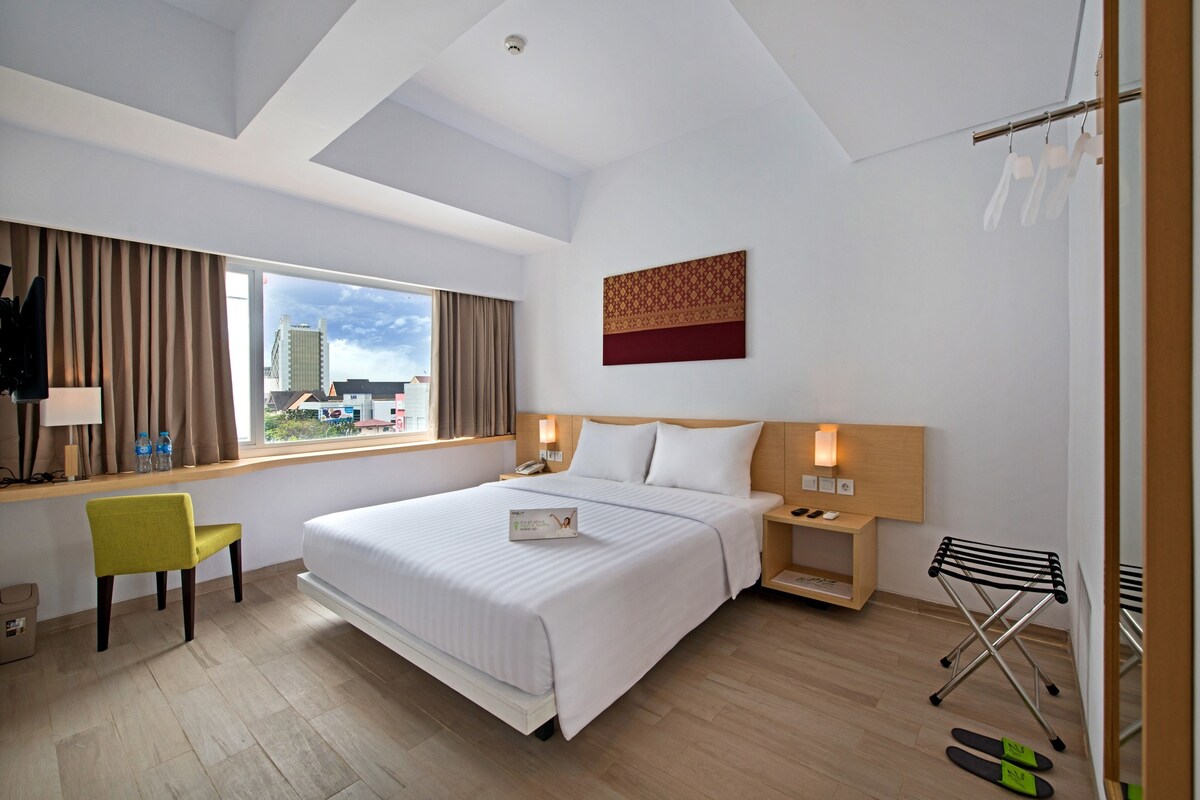 A modern hotel room with a large bed, a desk, a chair, a nightstand, a wall-mounted TV, and a window with curtains.