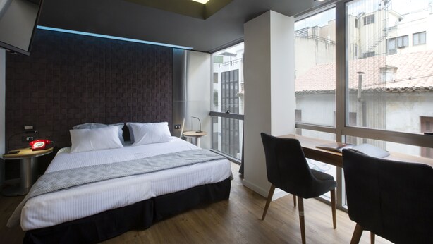 Quarto de hotel moderno com uma cama grande, uma escrivaninha e uma cadeira, televisão fixada na parede e grandes janelas com vista para a cidade.