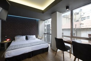 A modern hotel room with a large bed, a desk, and a chair, a TV mounted on the wall, and large windows offering a view of the city.