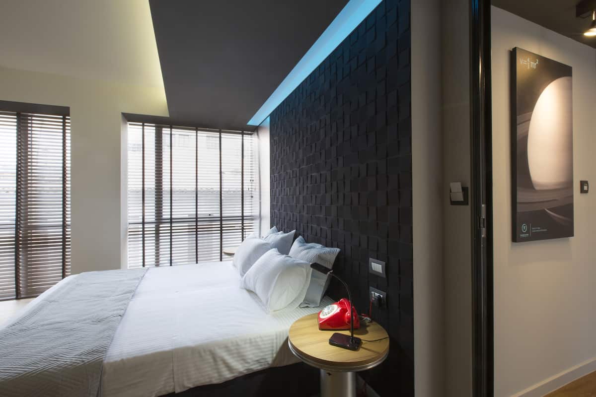 A modern hotel room with a bed, a round wooden nightstand, a red telephone, and a wall-mounted light.