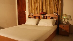Una cama bien tendida con sábanas blancas, cabecera de madera y una mesita de noche con una lámpara y un jarrón de flores.