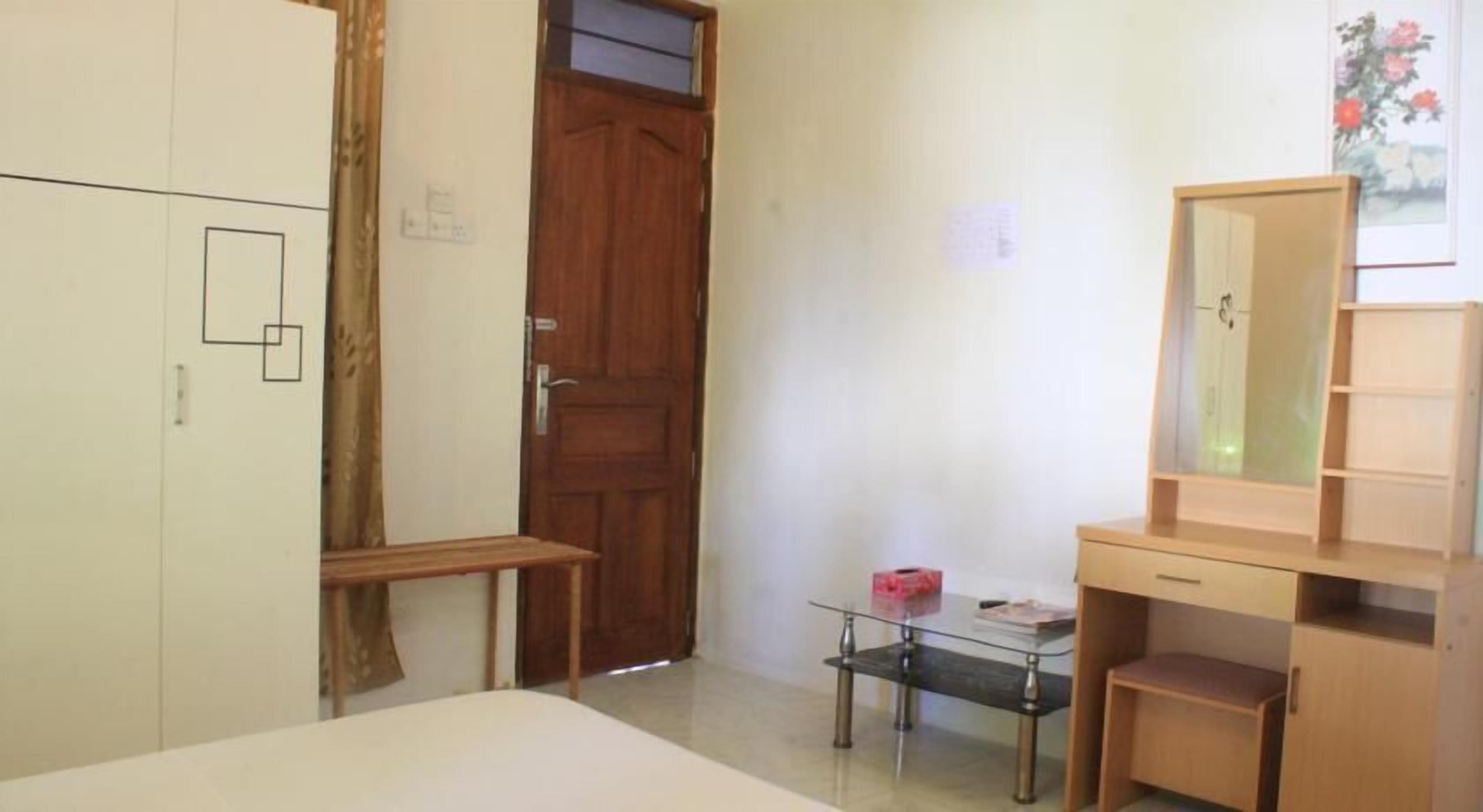 A room with a bed, a wooden wardrobe, a desk with a mirror, and a small table with a lamp.